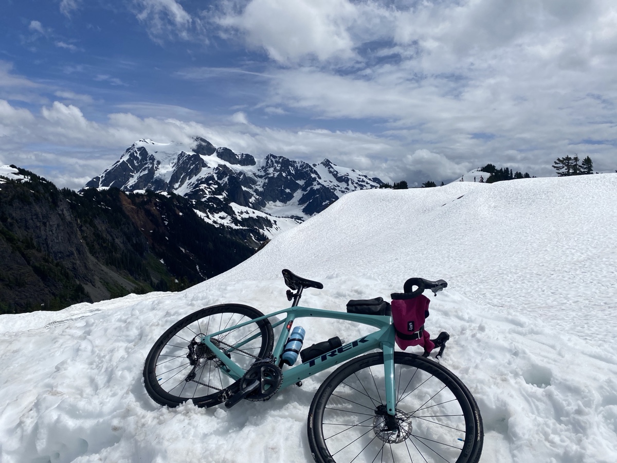 A turquoise road bike resting in snow beneath rugged mountain peaks and a cloudy blue sky.